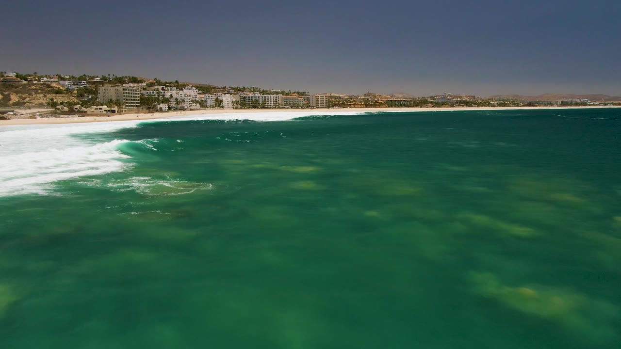 Pale underwater sand patches show through the green water at Costa Azul Beach while white surf breaks near the sandy shore and hillside buildings sit above it, with no visible people.