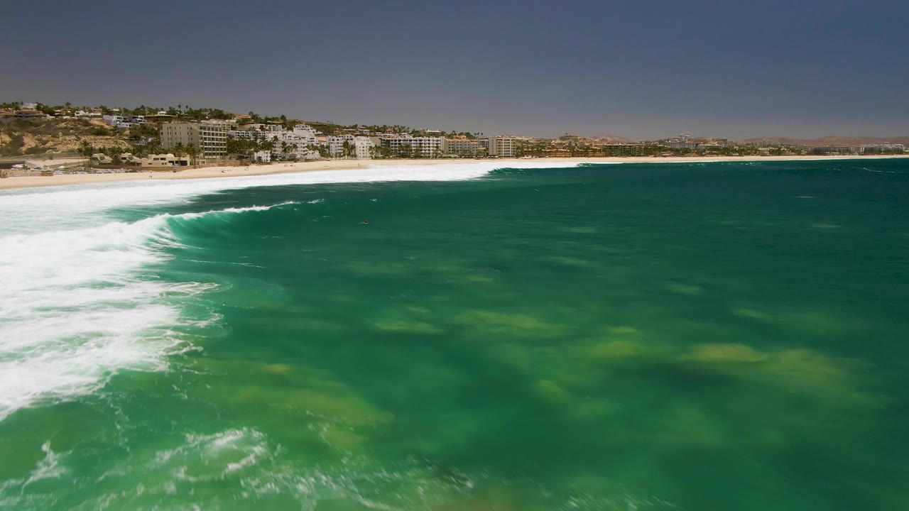 Aerial frame over Costa Azul Beach in Los Cabos showing white surf curling into green water, a sandy shoreline, hillside buildings, and no visible people.