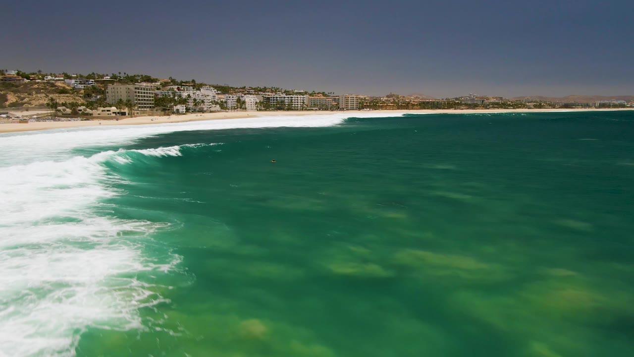Aerial view at Costa Azul Beach with a broader band of white foam crossing the green water below distant beachside buildings and no visible people.