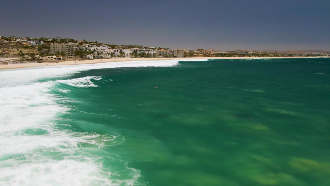 Costa Azul Beach aerial still with a curved white surf edge, clear green water in the foreground, sandy beach at left, distant buildings, and no visible people.