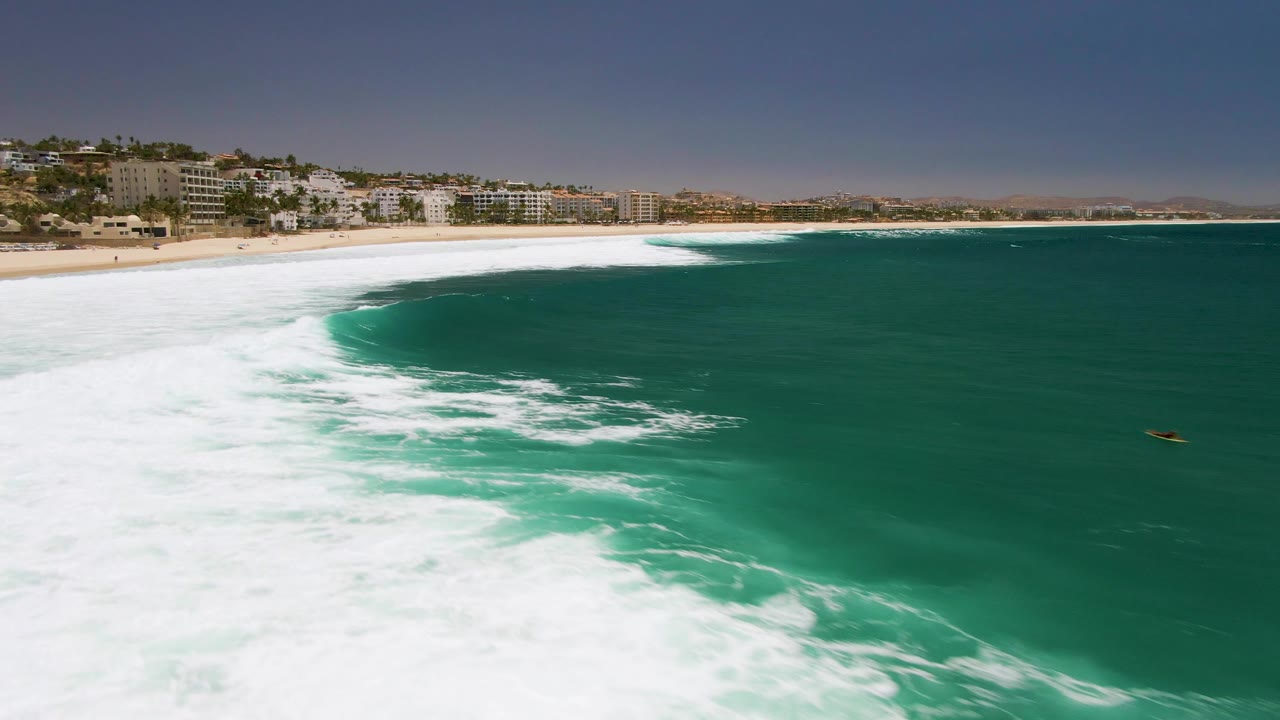 Aerial view of turquoise surf curving toward a sandy beach with coastal buildings beyond.