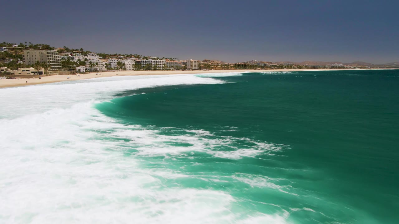 At Costa Azul Beach in Los Cabos, the white foam spreads farther across the turquoise ocean near the sand, with beachfront buildings and clear sky in the background and no visible people.