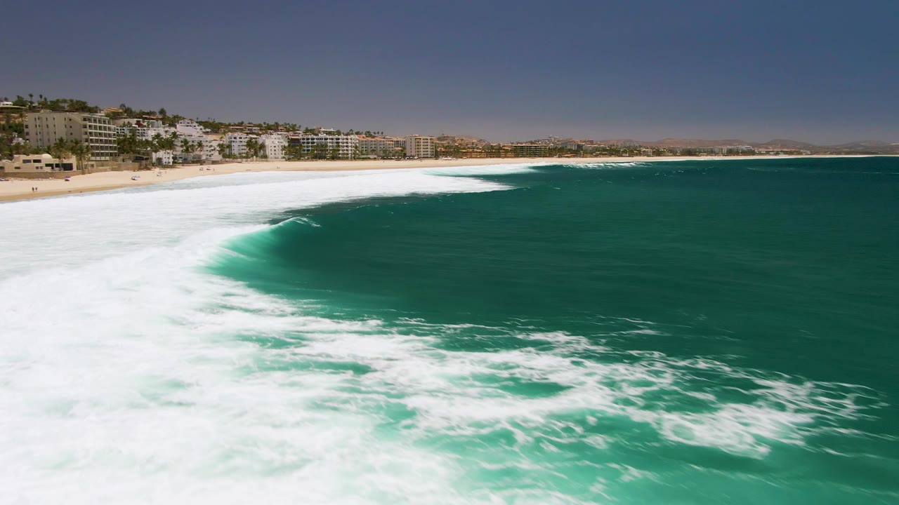 At Costa Azul Beach in Los Cabos, a curved wave crest pushes white foam across the turquoise water, with sand and coastal buildings visible along the upper left and no visible people.