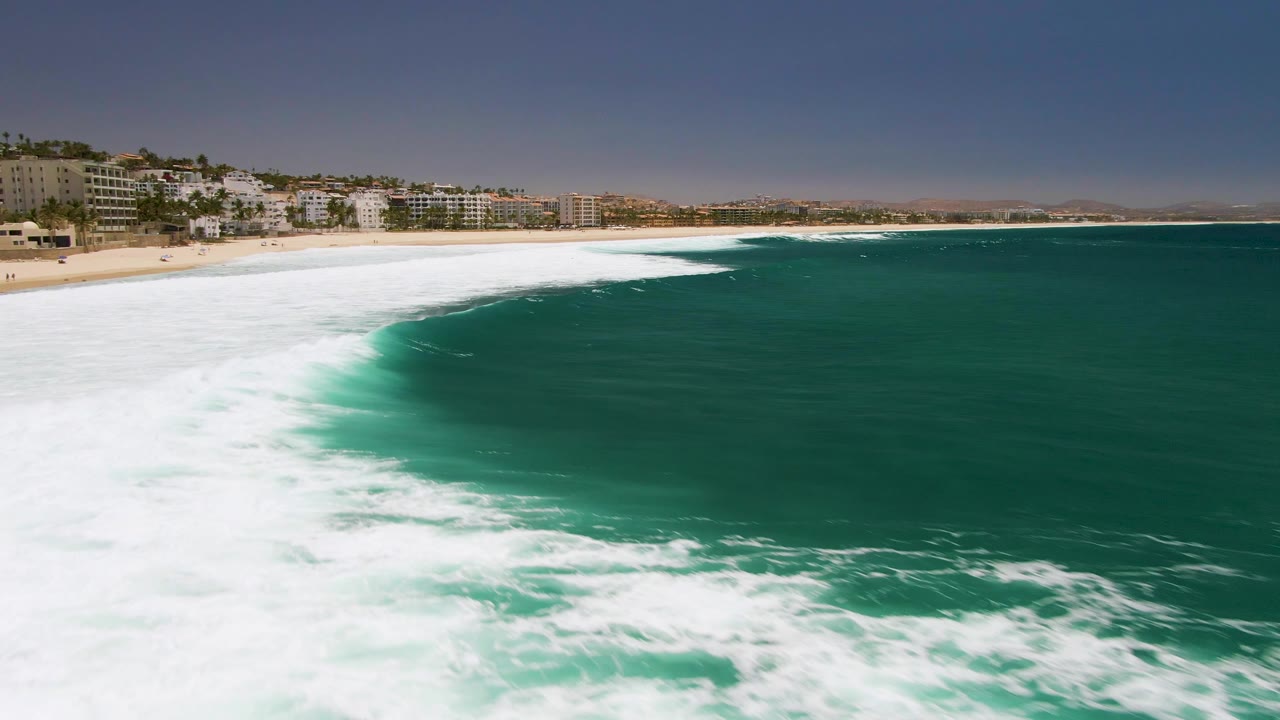 At Costa Azul Beach in Los Cabos, a wide white surf band cuts across deep turquoise water near the beach, with coastal buildings along the sand and no visible people.