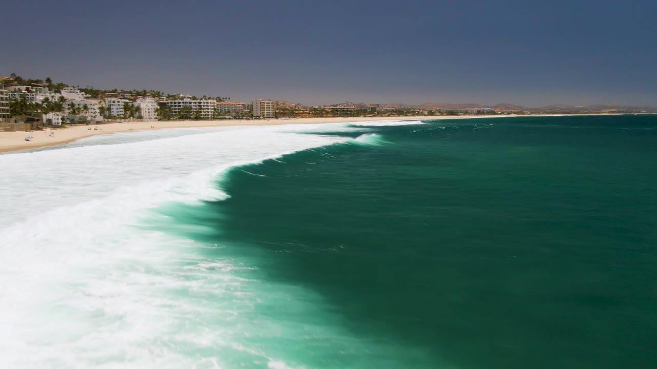At Costa Azul Beach in Los Cabos, green ocean water and a broad white surf band curve toward the sandy shore, with coastal buildings in the background and no visible people.