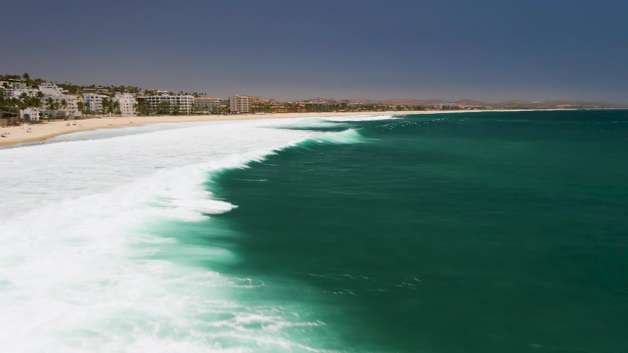 At 0:01, the foamy wave edge advances farther across the green water at Costa Azul Beach, with sand and low coastal buildings along the upper left and no visible people.