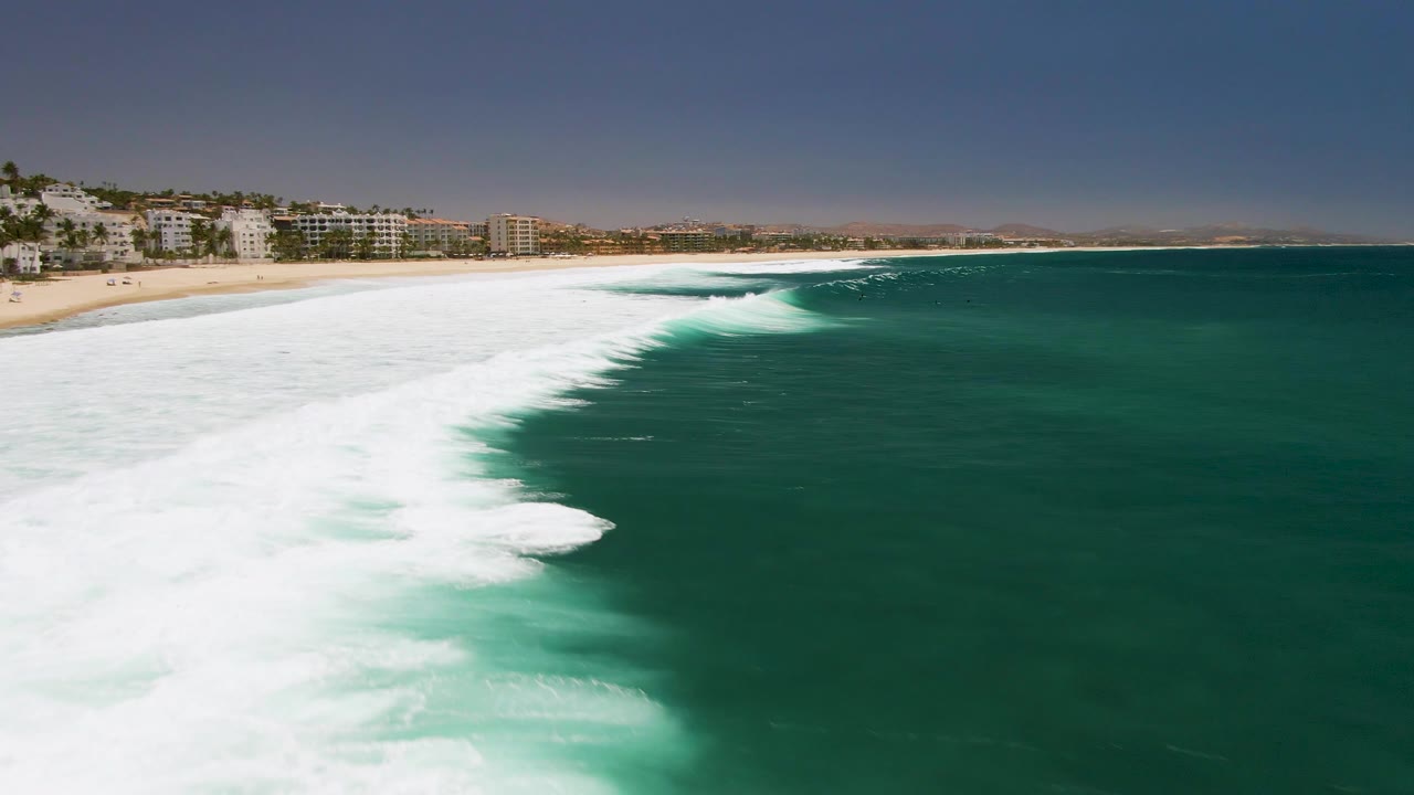 At Costa Azul Beach in Los Cabos, a long white breaker stretches diagonally from the beach into green ocean water, with coastal buildings on the left background and no visible people.