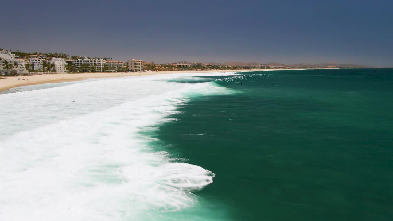 Foamy ocean water fills the left foreground as green water and sandy beach extend toward coastal buildings.