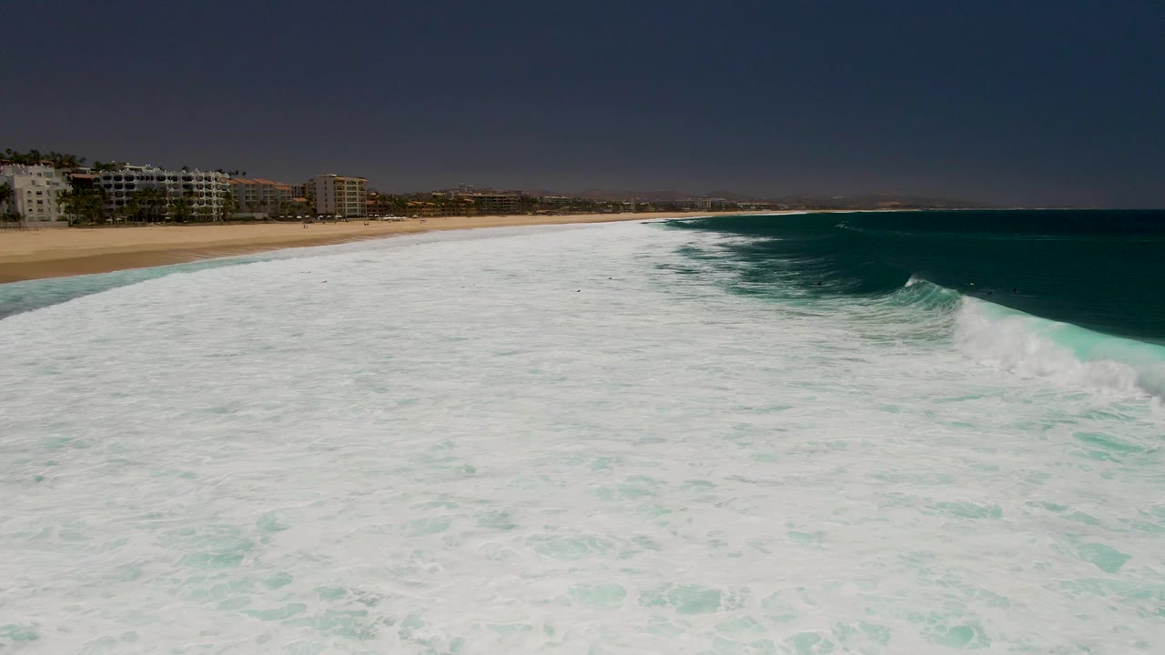 Aerial frame from Costa Azul Beach in Los Cabos showing broad white surf in the foreground, sand and beachfront buildings along the left, turquoise water to the right, and no visible people.