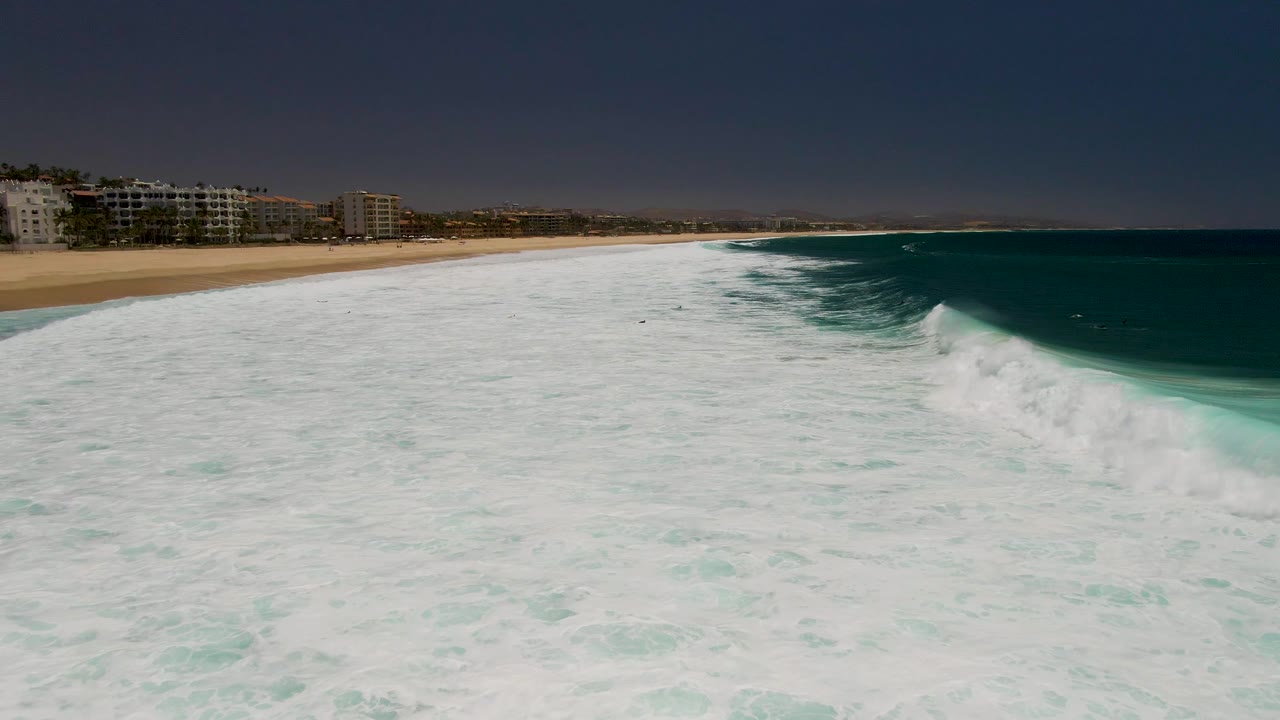 Aerial frame from Costa Azul Beach in Los Cabos with white surf covering most of the foreground, a curling turquoise wave on the right, beachfront buildings on the left, and no visible people.