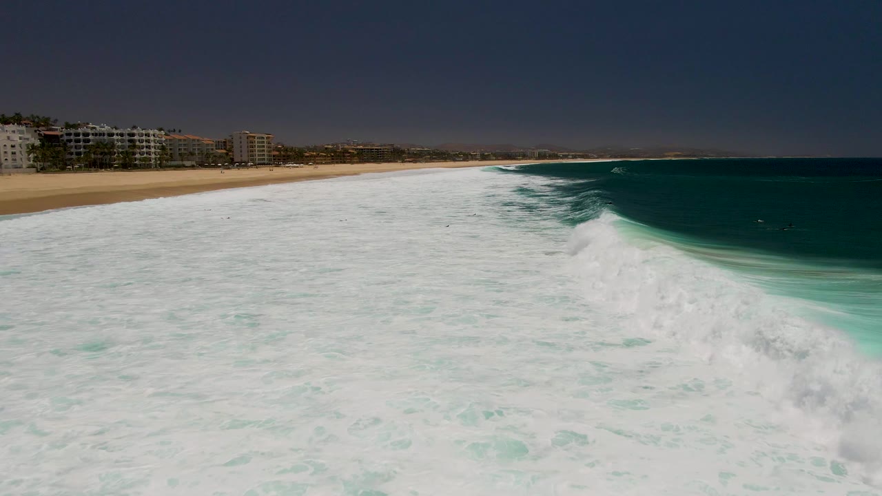 Aerial frame from Costa Azul Beach in Los Cabos showing a larger white breaking wave on the right, textured foam across the foreground, tan beach at left, distant buildings, and no visible people.