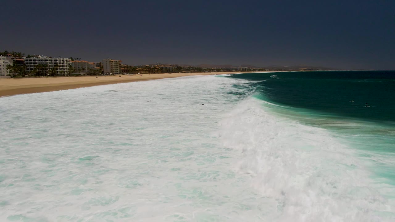 White surf fills the foreground beside a sandy beach and dark turquoise ocean.
