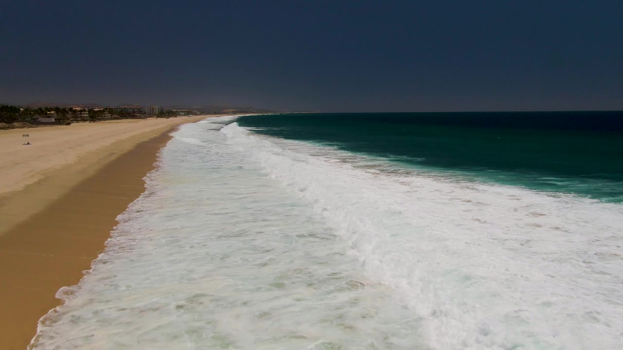 Aerial view at Costa Azul Beach in Los Cabos showing white surf spread across the shoreline, deep blue-green ocean to the right, tan sand to the left, and no visible people.