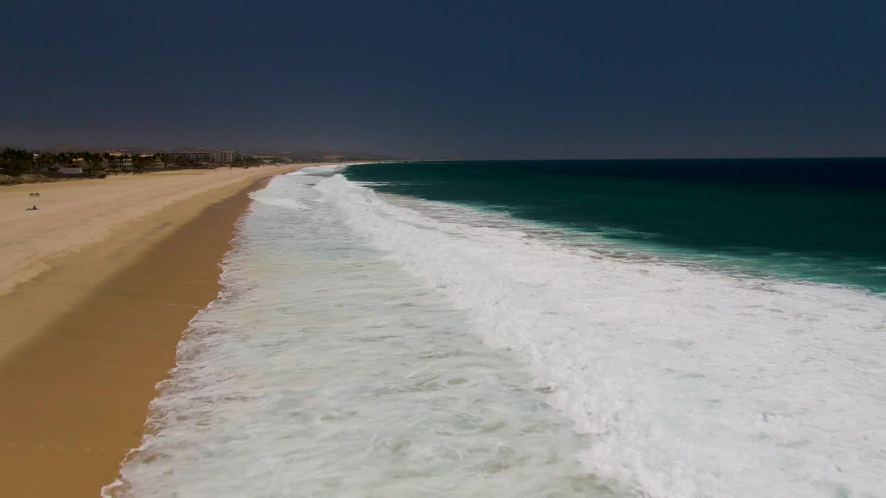 Foamy waves cover the shoreline with open ocean on the right and sand on the left.