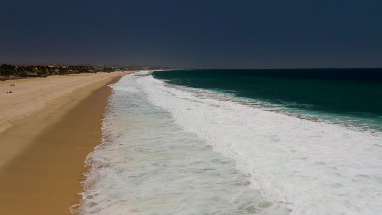 At 0:02, the Costa Azul Beach shoreline shows a curved edge of white breaking water beside tan sand, darker ocean beyond the foam, small distant structures, and no visible people.
