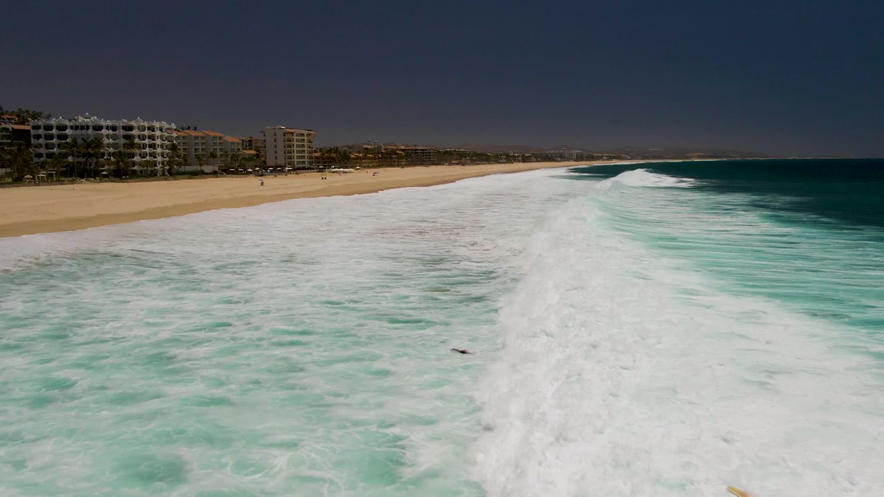 Aerial frame at Costa Azul Beach Los Cabos showing white surf filling the lower foreground, sandy beach and coastal buildings at left, turquoise ocean at right, and no visible people.