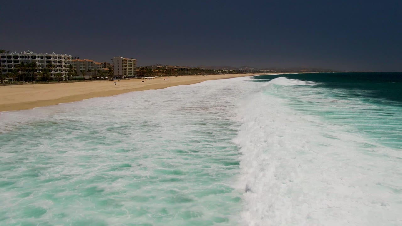 Foamy wave line running along a sandy beach with buildings in the distance.