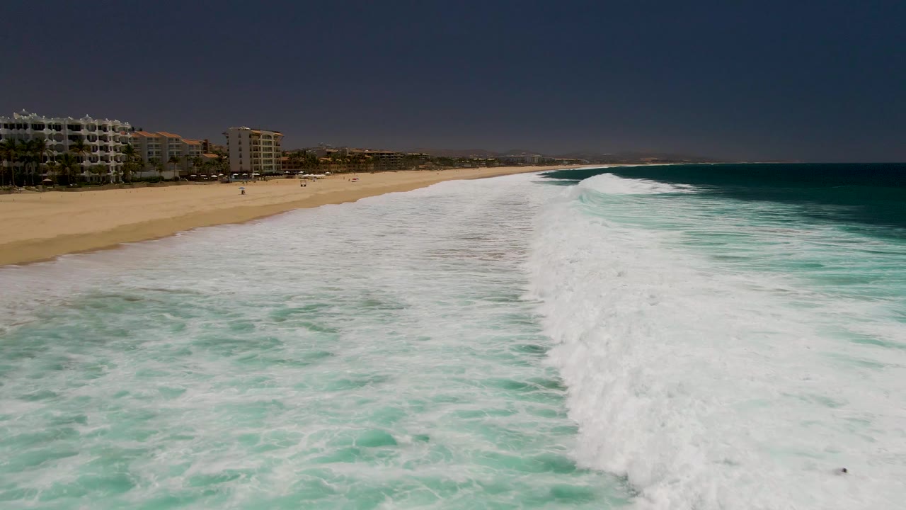 Aerial frame at Costa Azul Beach Los Cabos showing churned turquoise water in the foreground, a thick white surf break near center, sandy shore at left, background buildings, and no visible people.
