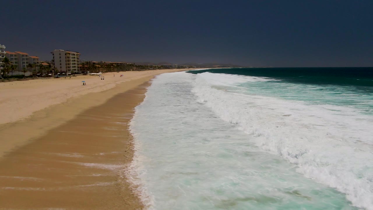 Aerial view at Costa Azul Beach in Los Cabos showing broad sand, white surf, turquoise water, dark sky, coastal buildings at left, and small distant people on the beach.