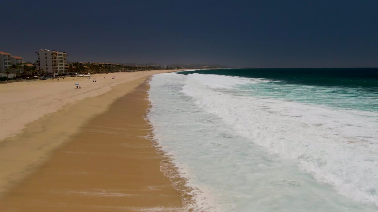 Foamy surf spreads along a sandy shoreline with buildings and small beach figures in the distance.