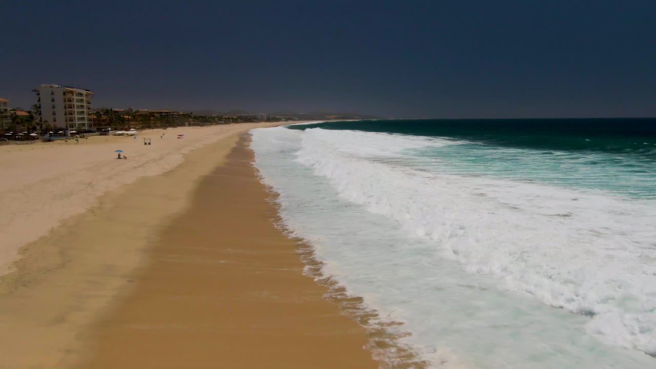 Aerial view at Costa Azul Beach in Los Cabos showing a longer stretch of tan sand, breaking white waves on the right, dark blue sky, coastal buildings, and tiny people on the beach.