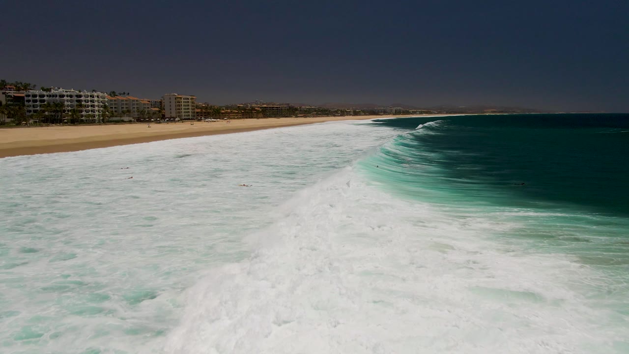 At Costa Azul Beach in Los Cabos, white ocean foam spreads across the foreground while a sandy beach, coastal buildings, and small swimmers sit in the distance.