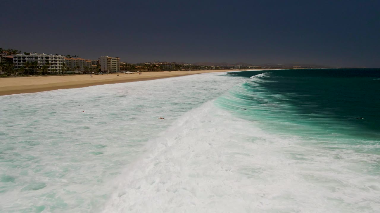 Whitewater and turquoise surf fill the frame beside a sandy shoreline and beach buildings.