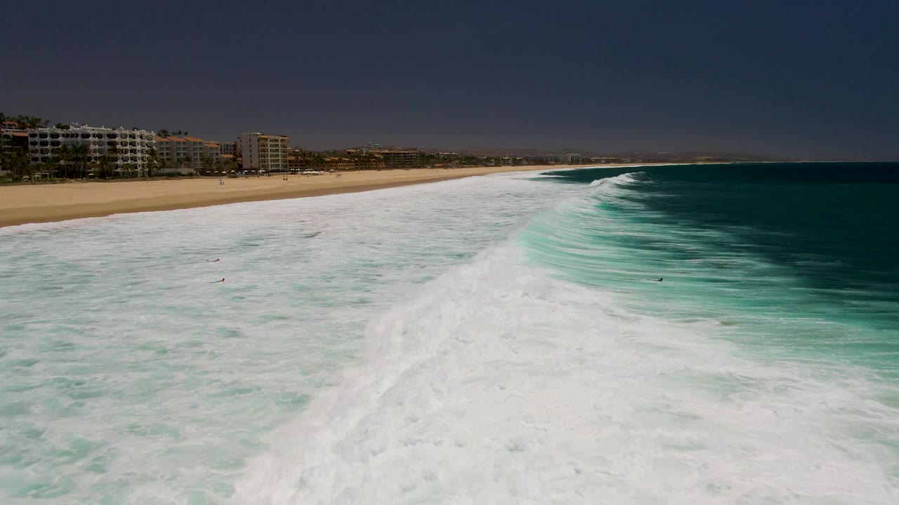 At Costa Azul Beach in Los Cabos, a broad curve of white foam cuts through turquoise water with small swimmers scattered below the sandy beach and coastal buildings.