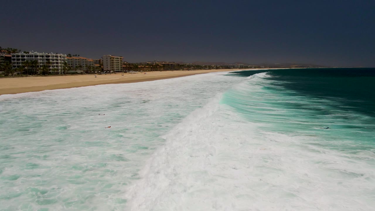 Wide aerial view of foamy waves beside a sandy beach with beachfront buildings.