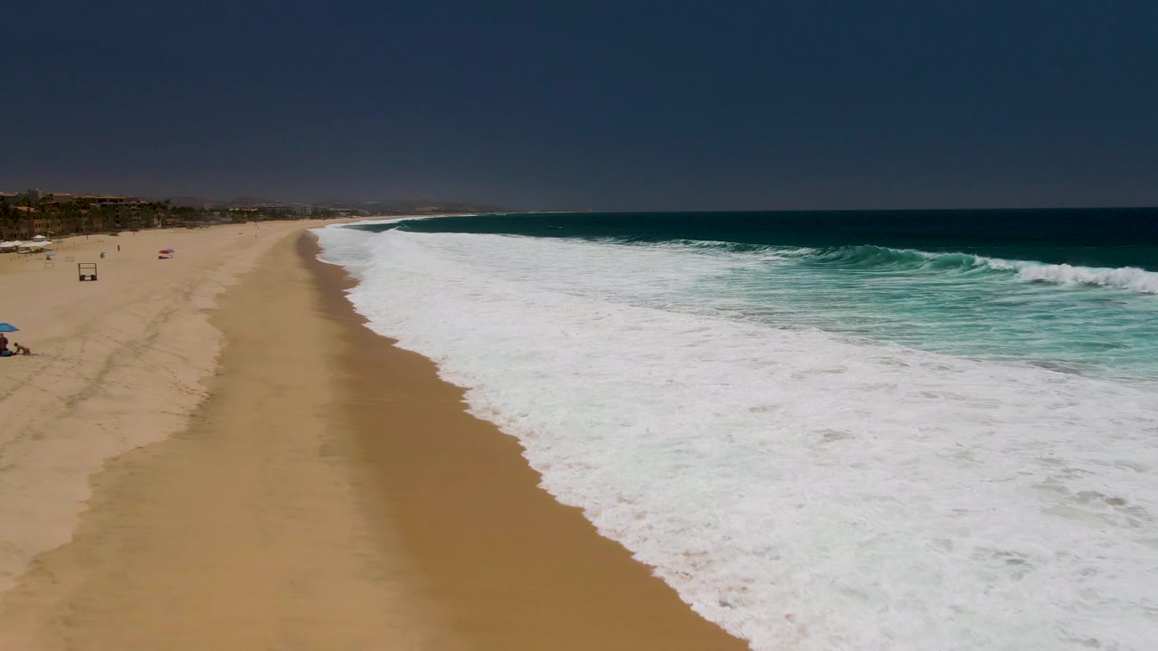 Aerial frame of Costa Azul Beach in Los Cabos with a wide sandy shore on the left, white surf rolling in from the right, turquoise water beyond the break, and tiny beachgoers near the far left edge.