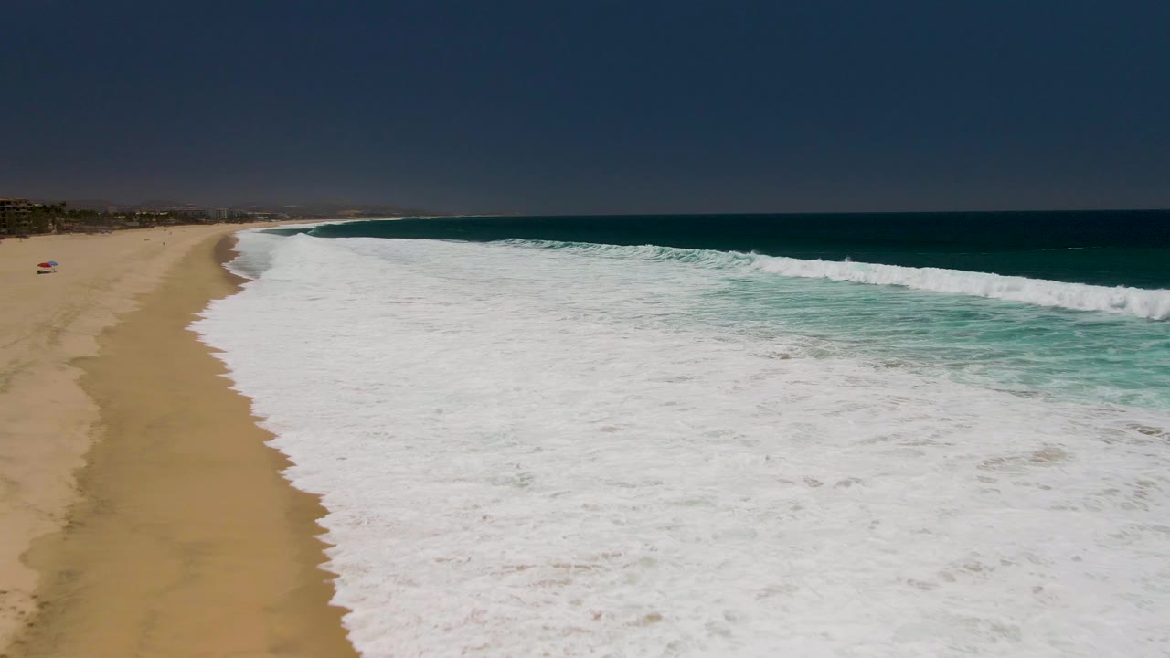 Aerial view of Costa Azul Beach with a narrow strip of sand at left, dense whitewater across the lower and right frame, turquoise water near the wave face, and a straight dark ocean horizon above.