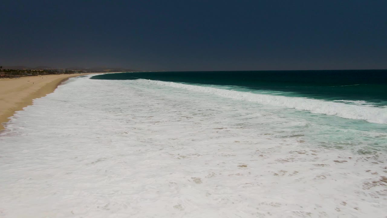 Aerial view of white surf rolling onto a sandy beach beside deep blue ocean.