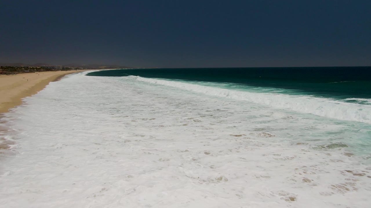 Aerial view at Costa Azul Beach with foamy water spreading wider across the lower frame, tan beach curving along the left side, and no visible people.