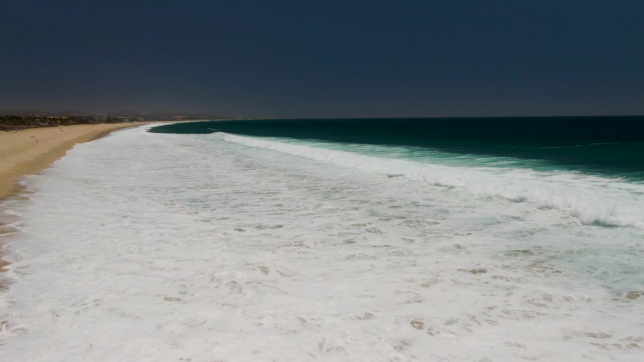 Costa Azul Beach shoreline from above, with dense white water across the foreground, a narrow strip of sand at left, dark blue ocean beyond, and no visible people.