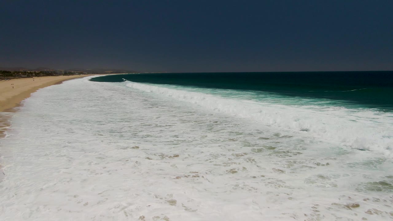 Aerial view over Costa Azul Beach as the surf line angles from the lower foreground toward the distant coast, with open ocean to the right and no visible people.