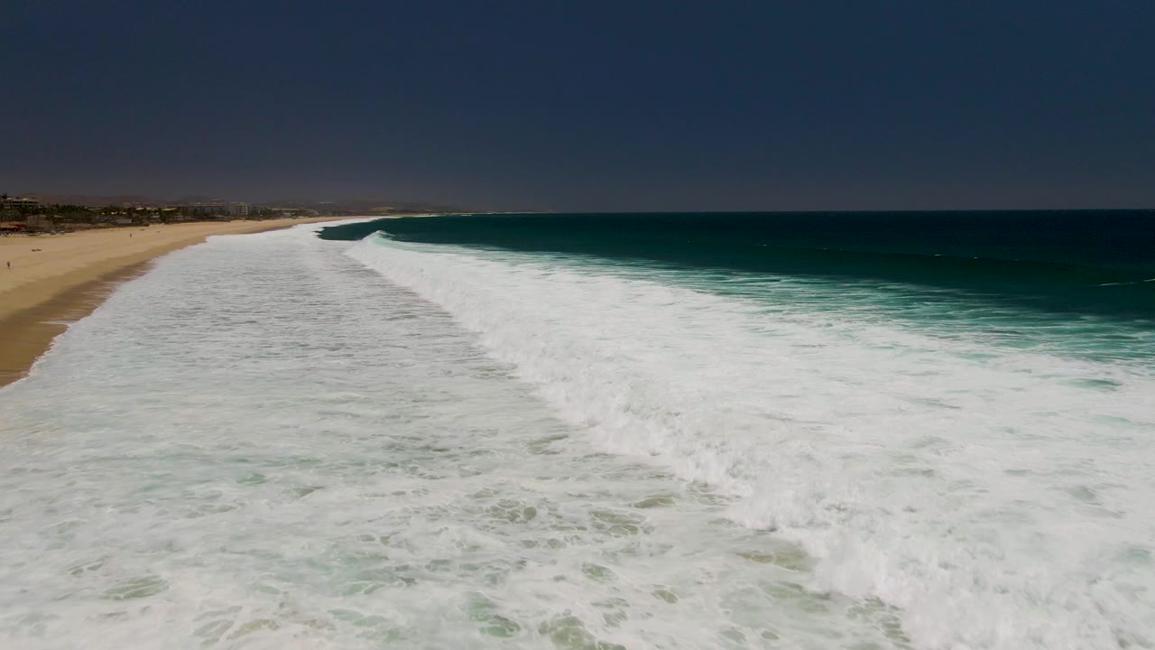 Aerial view at Costa Azul Beach in Los Cabos showing a wide band of white surf spreading toward tan sand, deep blue ocean to the right, and no visible people.