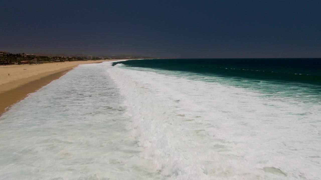 A long breaker at Costa Azul Beach runs diagonally along the sand, with a broad white foam field in front, blue ocean on the right, distant coastal development, and no visible people.