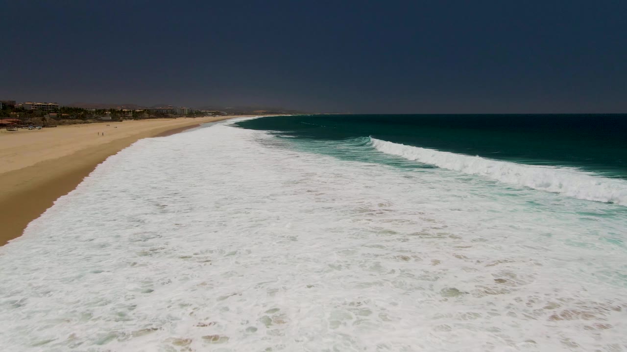 Aerial frame at Costa Azul Beach, Los Cabos, showing a broad sheet of white foam beside tan sand on the left and deep blue-green ocean on the right, with no visible people.