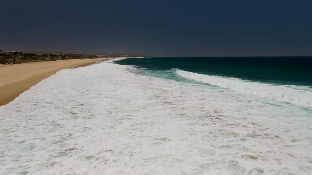 Foamy wave spreading across water beside a long sandy shoreline.