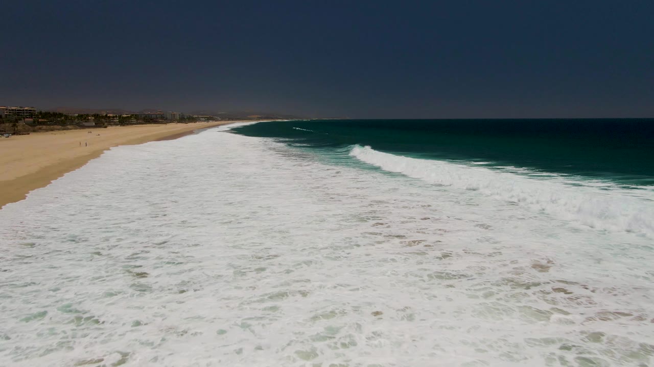 Costa Azul Beach appears from above as a breaking surf line rolls across the right side, white water fills the foreground, and the tan beach remains visible along the left, with no visible people.