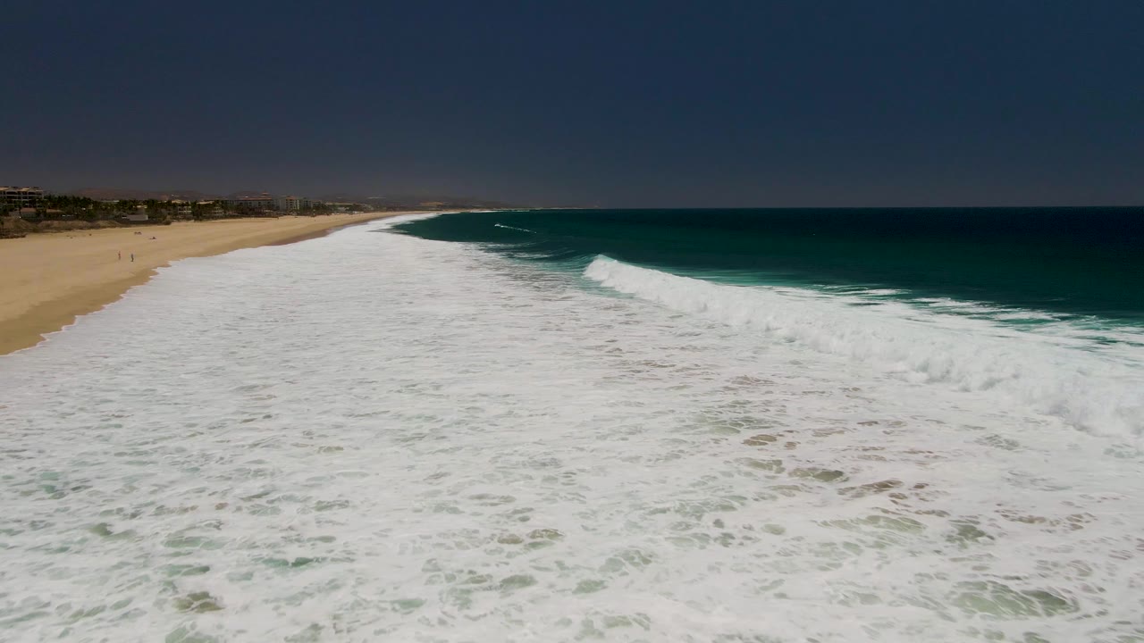 Wide aerial beach view with dense white surf and ocean horizon.