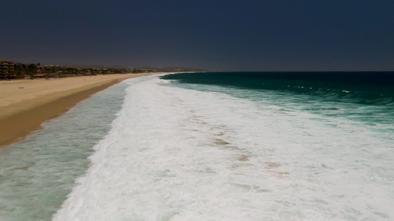 Aerial view at Costa Azul Beach in Los Cabos with white surf spread across the foreground, tan sand on the left, blue ocean on the right, and no visible people.