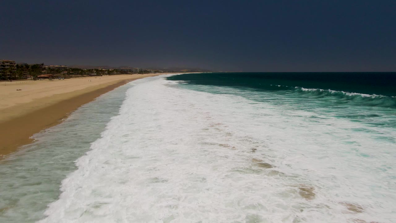 At Costa Azul Beach in Los Cabos, the foamy wave front extends farther across the center of the frame with sand on the left, darker ocean water on the right, and no visible people.