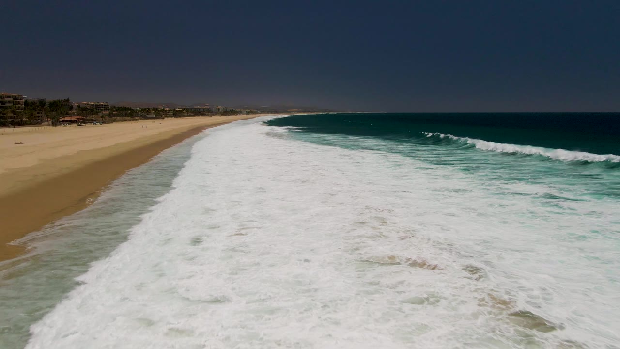 Aerial frame from Costa Azul Beach in Los Cabos with a wide band of white foam across the lower frame, open sand at left, deep blue water at right, and no visible people.