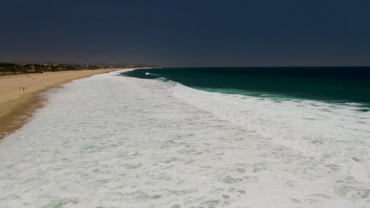 Aerial frame of Costa Azul Beach in Los Cabos with white foam covering the foreground, turquoise water to the right, sand to the left, and tiny beachgoers visible near the shore.