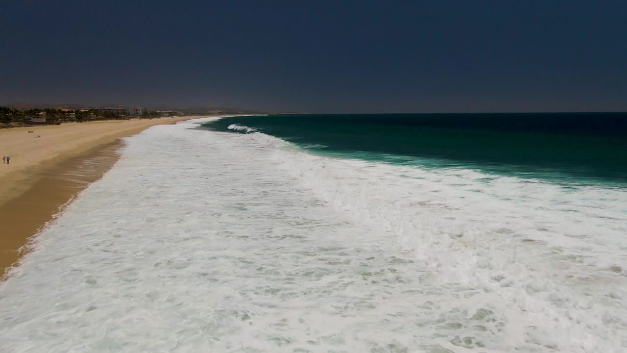 Aerial frame of Costa Azul Beach with dense white foam across the foreground, a clean dark blue horizon, turquoise water near the break, and tiny figures on the sand at left.