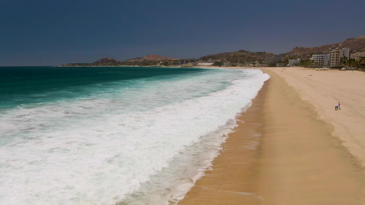 Aerial frame from Costa Azul Beach in Los Cabos, Mexico, showing white surf sweeping onto sand, deep blue ocean at left, coastal hills and buildings in the distance, and a tiny beach figure near the right edge.