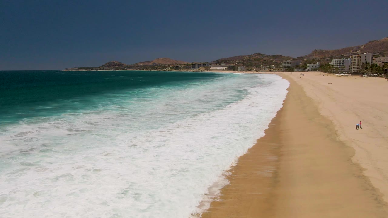 Aerial view over Costa Azul Beach with a broad white foam band curving toward wet sand, darker blue water offshore, distant hills, beachfront buildings, and a small beach figure at the far right.