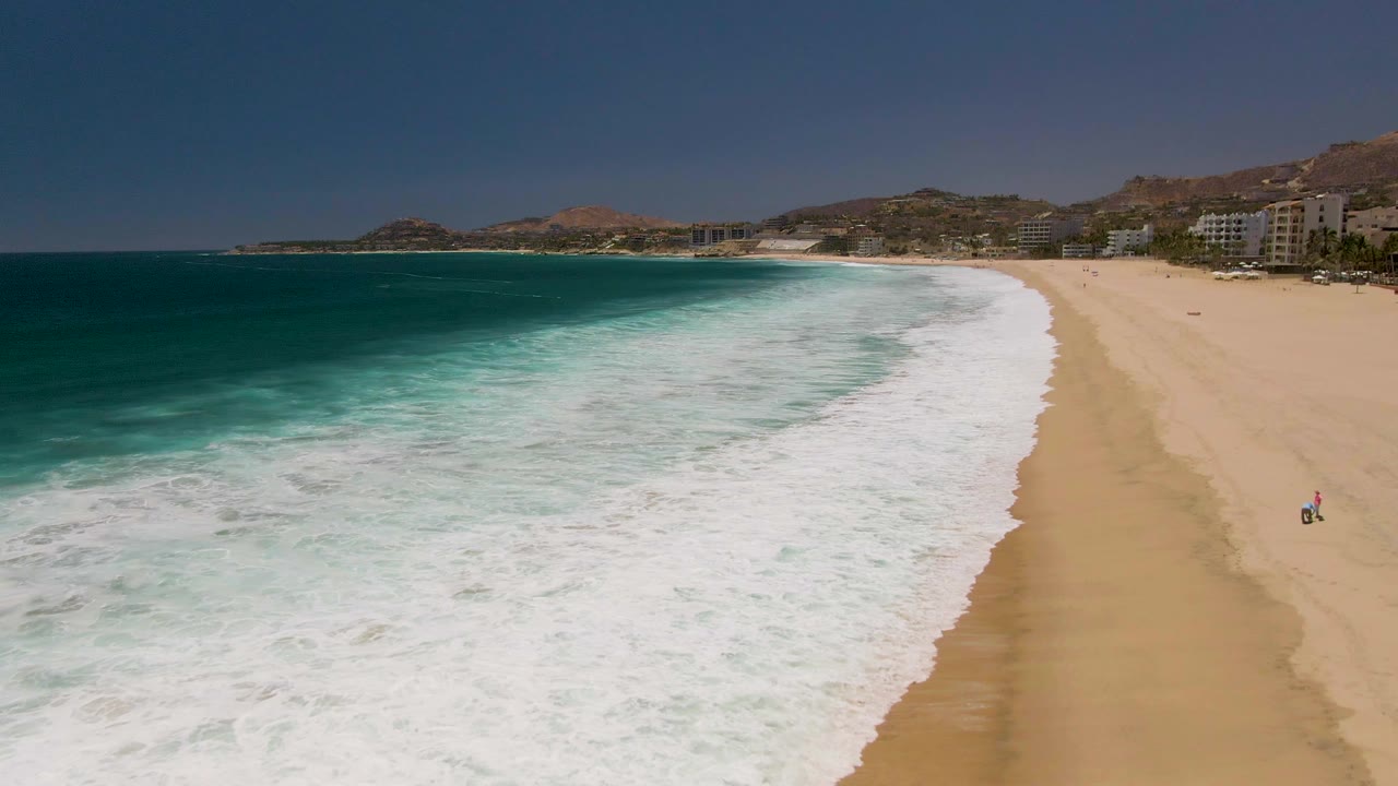 Costa Azul Beach appears from above with the surf spreading across the lower center, a long strip of tan sand on the right, blue ocean on the left, and small coastal buildings below dry hills.