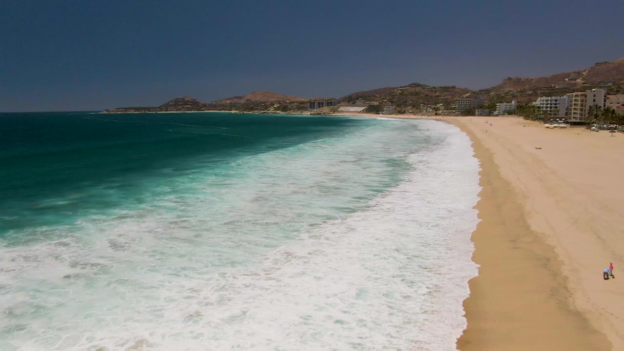 Aerial beach view with waves washing across sand and coastal buildings in the background.
